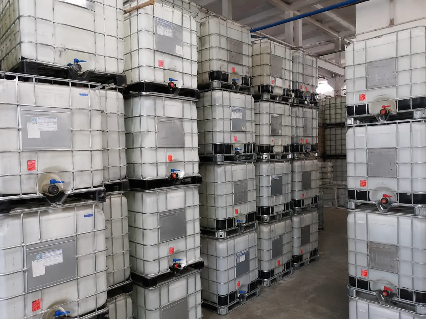 Reconditioned IBC totes stacked three high along a warehouse aisle, cages and discharge valves visible