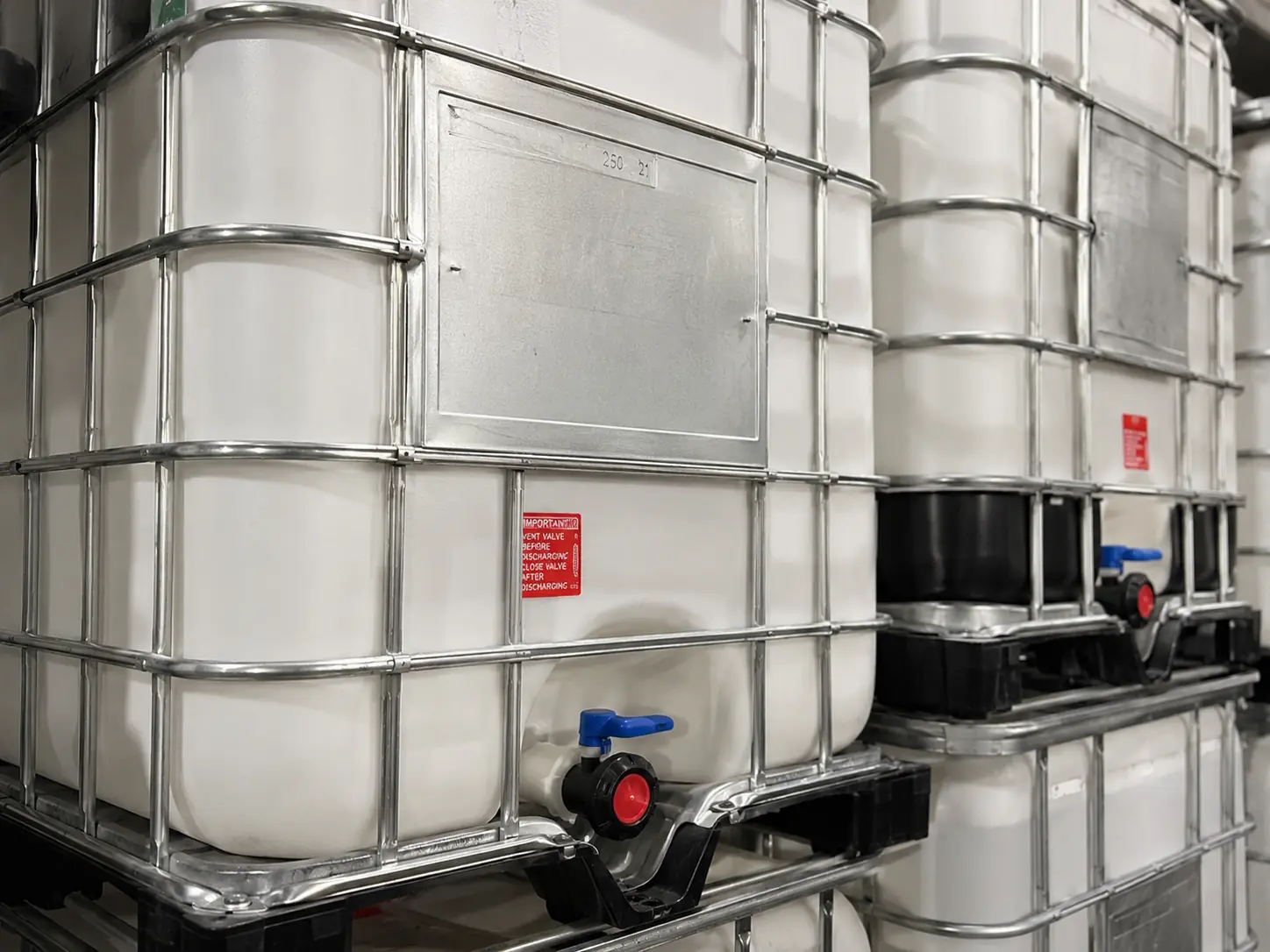 Close-up of a caged IBC tote showing the UN data plate, the two-inch discharge valve and the galvanised steel cage
